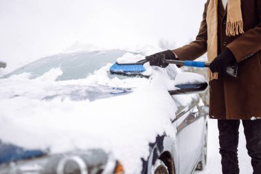 Palto ve eşarplı şık bir adam kış kar yağarken arabasındaki karları temizler. Mevsimlik konsept, hava durumu, insanlar, ulaşım.
