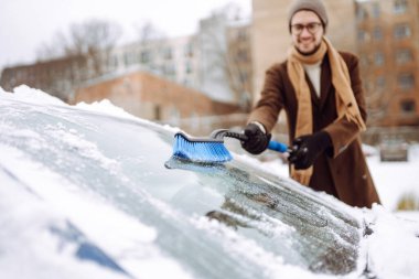 Palto ve eşarplı şık bir adam kış kar yağarken arabasındaki karları temizler. Mevsimlik konsept, hava durumu, insanlar, ulaşım.