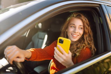 Bir kadın sürücü arabada akıllı telefon kullanır. Sürücü koltuğundaki mutlu kadın yön bulma aracıyla hareket ediyor. Seyahat, teknoloji, internet kavramı. Boş zaman aktiviteleri.