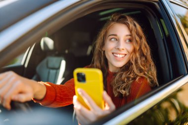 Bir kadın sürücü arabada akıllı telefon kullanır. Sürücü koltuğundaki mutlu kadın yön bulma aracıyla hareket ediyor. Seyahat, teknoloji, internet kavramı. Boş zaman aktiviteleri.