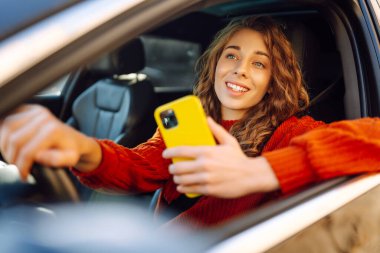Bir kadın sürücü arabada akıllı telefon kullanır. Sürücü koltuğundaki mutlu kadın yön bulma aracıyla hareket ediyor. Seyahat, teknoloji, internet kavramı. Boş zaman aktiviteleri.