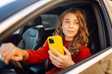 Bir kadın sürücü arabada akıllı telefon kullanır. Sürücü koltuğundaki mutlu kadın yön bulma aracıyla hareket ediyor. Seyahat, teknoloji, internet kavramı. Boş zaman aktiviteleri.