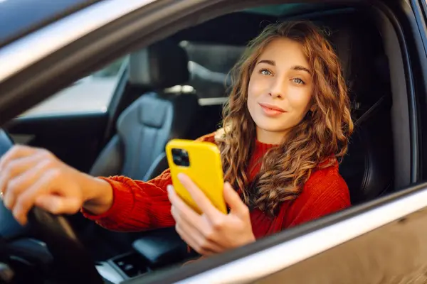 A woman driver uses a smartphone in the car. Happy woman in the driver\'s seat using navigation to move around. Concept of travel, technology, internet. Leisure activities.
