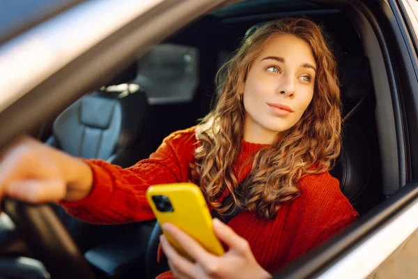 A woman driver uses a smartphone in the car. Happy woman in the driver\'s seat using navigation to move around. Concept of travel, technology, internet. Leisure activities.