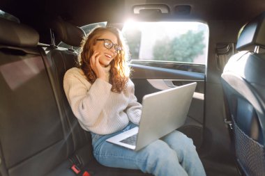 Arabanın arka koltuğunda dizüstü bilgisayarı olan gözlüklü genç bir kadın. İş kadını arabada dizüstü bilgisayarla çalışıyor. Serbest meslek, iş kavramı.