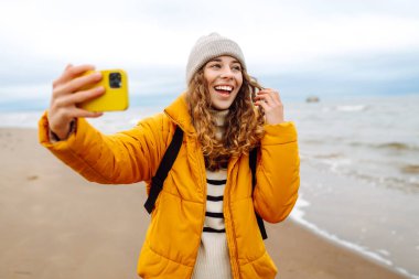 Deniz kıyısında elinde telefon olan parlak ceketli mutlu bir kadın. Genç turist kadın deniz kenarında selfie ya da blog çekiyor, deniz manzarasının tadını çıkarıyor. Seyahatler, hafta sonu.
