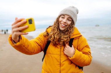 Deniz kıyısında elinde telefon olan parlak ceketli mutlu bir kadın. Genç turist kadın deniz kenarında selfie ya da blog çekiyor, deniz manzarasının tadını çıkarıyor. Seyahatler, hafta sonu.