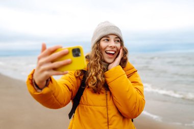 Deniz kıyısında elinde telefon olan parlak ceketli mutlu bir kadın. Genç turist kadın deniz kenarında selfie ya da blog çekiyor, deniz manzarasının tadını çıkarıyor. Seyahatler, hafta sonu.