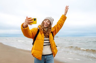 Deniz kıyısında elinde telefon olan parlak ceketli mutlu bir kadın. Genç turist kadın deniz kenarında selfie ya da blog çekiyor, deniz manzarasının tadını çıkarıyor. Seyahatler, hafta sonu.