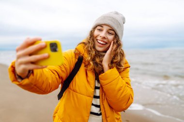 Deniz kıyısında elinde telefon olan parlak ceketli mutlu bir kadın. Genç turist kadın deniz kenarında selfie ya da blog çekiyor, deniz manzarasının tadını çıkarıyor. Seyahatler, hafta sonu.
