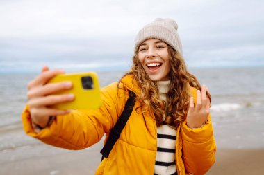 Deniz kıyısında elinde telefon olan parlak ceketli mutlu bir kadın. Genç turist kadın deniz kenarında selfie ya da blog çekiyor, deniz manzarasının tadını çıkarıyor. Seyahatler, hafta sonu.