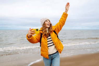 Deniz kıyısında elinde telefon olan parlak ceketli mutlu bir kadın. Genç turist kadın deniz kenarında selfie ya da blog çekiyor, deniz manzarasının tadını çıkarıyor. Seyahatler, hafta sonu.