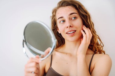 Dermatoloji. Güzel bir kadın aynaya bakıyor, elleriyle yüzüne dokunuyor, sivilceleri ve pigmentasyonu inceliyor. Kozmetik bakım. Alerjik reaksiyon. Sağlık konsepti, kozmetoloji.