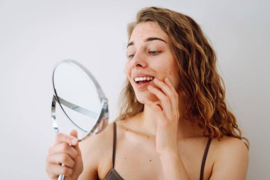 Dermatoloji. Güzel bir kadın aynaya bakıyor, elleriyle yüzüne dokunuyor, sivilceleri ve pigmentasyonu inceliyor. Kozmetik bakım. Alerjik reaksiyon. Sağlık konsepti, kozmetoloji.