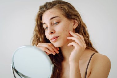 Dermatoloji. Güzel bir kadın aynaya bakıyor, elleriyle yüzüne dokunuyor, sivilceleri ve pigmentasyonu inceliyor. Kozmetik bakım. Alerjik reaksiyon. Sağlık konsepti, kozmetoloji.