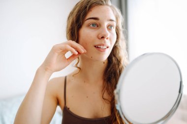 Dermatoloji. Güzel bir kadın aynaya bakıyor, elleriyle yüzüne dokunuyor, sivilceleri ve pigmentasyonu inceliyor. Kozmetik bakım. Alerjik reaksiyon. Sağlık konsepti, kozmetoloji.