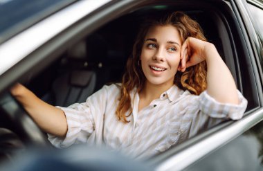 Arabasını süren gülümseyen kadın. Sıradan giysiler içinde araba süren şık bir kadının portresi. Araba yolculuğu kavramı.