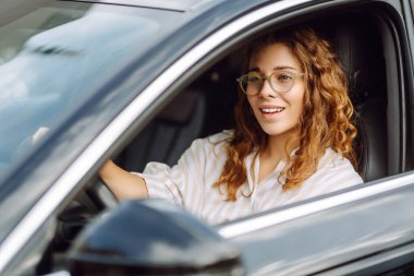 Arabasını süren gülümseyen kadın. Sıradan giysiler içinde araba süren şık bir kadının portresi. Araba yolculuğu kavramı.