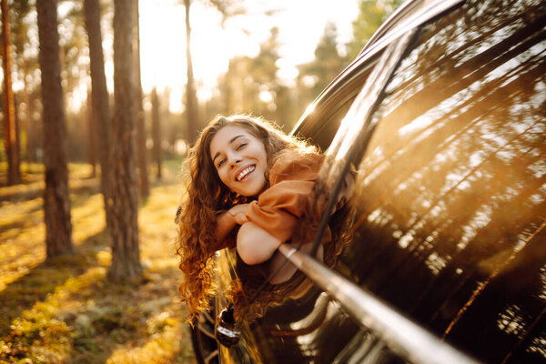 Happy woman in a car catches the wind from the car window, enjoying nature. Young woman on a car trip. Travel, adventure concept. Active lifestyle.