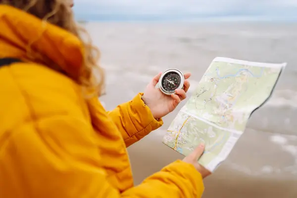 A young woman traveler with a compass and a map in her hands, reading a map looking for directions on the beach near the sea. Adventure, vacation concept. Active lifestyle.
