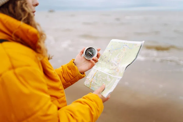 A young woman traveler with a compass and a map in her hands, reading a map looking for directions on the beach near the sea. Adventure, vacation concept. Active lifestyle.