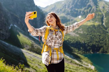 Sarı sırt çantalı ve elinde telefon olan mutlu bir bayan gezgin dağ manzaralarının arka planında selfie çekip blog yazıyor. Macera, tatil konsepti.