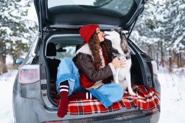 Kış tatilinde arabayla seyahat ederken, köpeğiyle karlı bir yolda bir arabanın bagajında piknik yapan mutlu bir kadın. Seyahat kavramı.