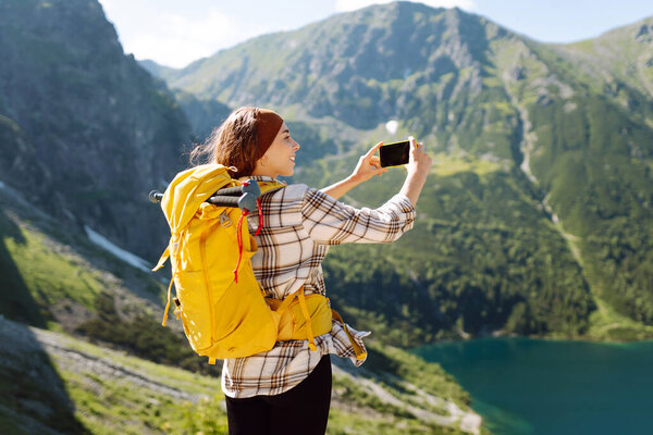 Happy female traveler with a yellow backpack and a phone in her hands takes a selfie and blogs against the backdrop of mountain landscapes. Adventure, vacation concept.