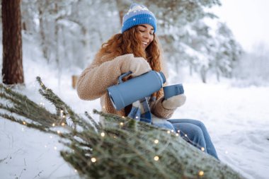 Doğada sarılı Noel ağacı olan mutlu bir kadın termostan sıcak bir içecek içer. Genç bir kadın kış havasının tadını çıkarıyor. Kış tatili kavramı, mutluluk..