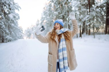 Parlak şapkalı ve atkılı mutlu kadın elinde telefonla güneşli bir kış ormanında selfie çekiyor. Seyahat konsepti, teknoloji, eğlence.