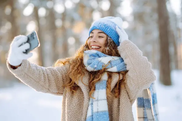 Parlak şapkalı ve atkılı mutlu kadın elinde telefonla güneşli bir kış ormanında selfie çekiyor. Seyahat konsepti, teknoloji, eğlence.