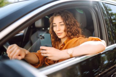 Araba kullanırken elinde telefonla, günlük kıyafetlerle dolaşan genç bir kadın. Mutlu kadın araba kiralamak için mobil uygulama kullanıyor. Teknoloji kavramı, ulaşım.