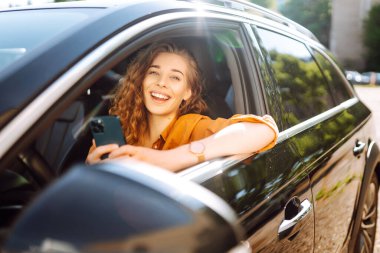 Araba kullanırken elinde telefonla, günlük kıyafetlerle dolaşan genç bir kadın. Mutlu kadın araba kiralamak için mobil uygulama kullanıyor. Teknoloji kavramı, ulaşım.