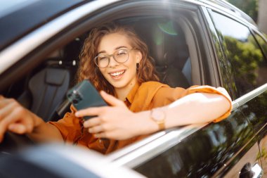 Araba kullanırken elinde telefonla, günlük kıyafetlerle dolaşan genç bir kadın. Mutlu kadın araba kiralamak için mobil uygulama kullanıyor. Teknoloji kavramı, ulaşım.