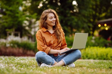 Yeşil çimenlikte dizüstü bilgisayarı olan sıradan giyinmiş güzel bir kadın. Dışarıda dizüstü bilgisayarda çalışan mutlu serbest çalışan kadın. Eğitim ve serbest meslek kavramı.