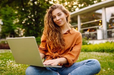 Yeşil çimenlikte dizüstü bilgisayarı olan sıradan giyinmiş güzel bir kadın. Dışarıda dizüstü bilgisayarda çalışan mutlu serbest çalışan kadın. Eğitim ve serbest meslek kavramı.