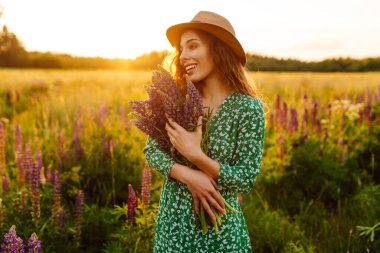 Lupin dolu bir tarlada elinde bir buket çiçekle hasır şapkalı şık bir kadın. Gün batımında yürürken doğanın tadını çıkaran genç bir kadın. Doğa konsepti.