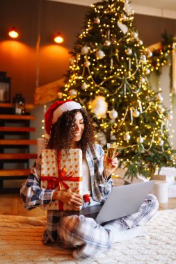 Güzel bir kadın Noel ağacının yanında hediyeli bir dizüstü bilgisayar kullanıyor. Genç bir kadın evdeki Noel ağacının yanında online alışveriş yapıyor. Tatil konsepti, sürpriz..