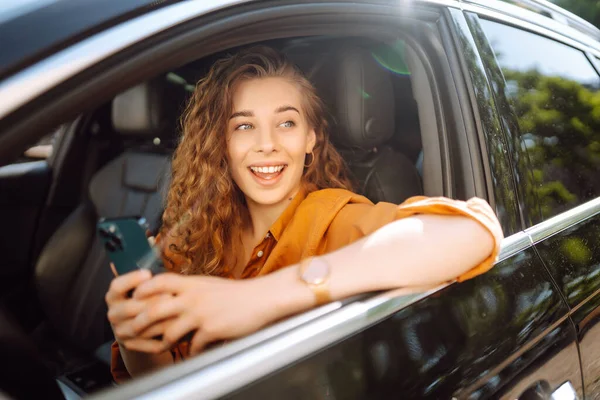 Young woman in casual clothes with a phone in her hands while driving a car. Happy woman uses a mobile application to rent a car. Concept of technology, transport.