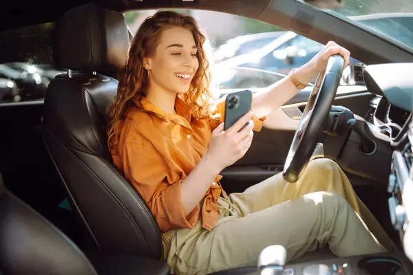 Young woman in casual clothes with a phone in her hands while driving a car. Happy woman uses a mobile application to rent a car. Concept of technology, transport.