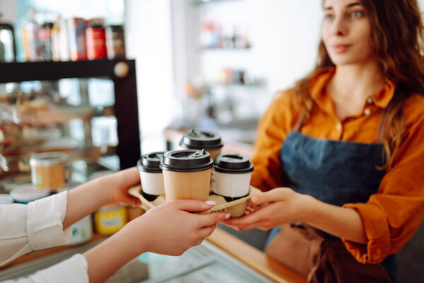 Close-up of a female barista's hands passing prepared drinks to go. A young woman owner of a coffee shop behind the bar prepares coffee. Concept of orders to go.