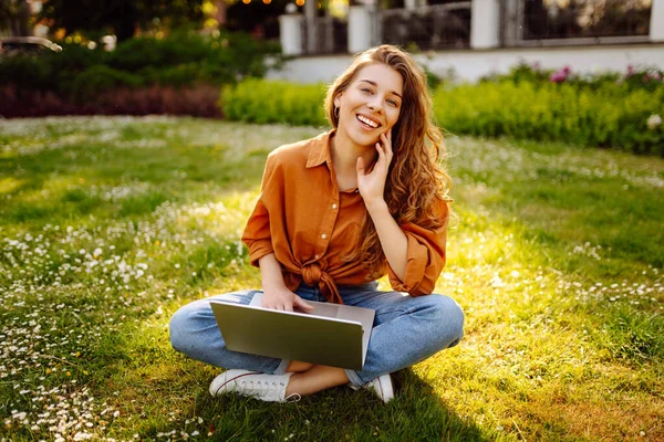Beautiful woman in casual clothes on a green lawn with a laptop. Happy freelancer woman working on laptop outdoors. Education and freelancing concept.