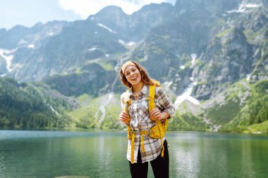 Yüksek bir dağ gölünde parlak bir sırt çantasıyla genç bir kadın gezgin. Gezginlerin görüntüsü. Macera, tatil konsepti. Etkin yaşam biçimi.