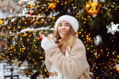 Genç bir kadın Noel pazarında sıcak bir içecek içiyor. Akşamları festival ışıklarıyla süsleniyor. Tatil konsepti, hafta sonları, rahatlama..