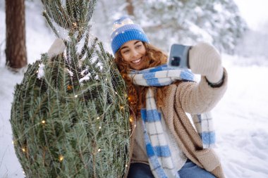 Mavi şapkalı, eşarplı, Noel ağacı ve telefonu olan mutlu bir kadın. Telefonlu genç bir kadın ve çelenkli bir Noel ağacı güzel kış havasının tadını çıkarıyor..