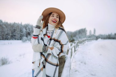 Kırmızı dudaklı, şapkalı ve açık renk eşarplı güzel bir kadın karlı bir ormanda kış gününün tadını çıkarıyor. Açık havada poz veren mutlu bayan turist. Noel.
