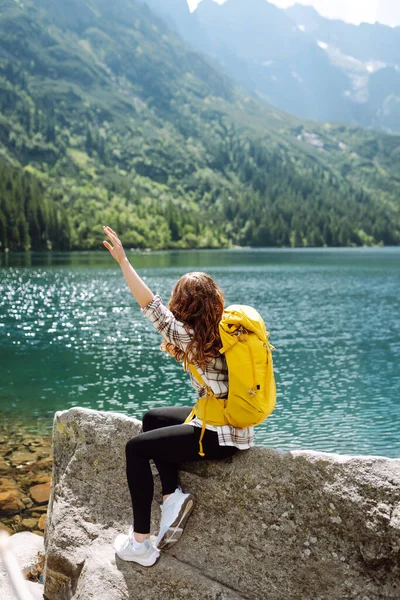 Yüksek bir dağ gölünde parlak bir sırt çantasıyla genç bir kadın gezgin. Gezginlerin görüntüsü. Macera, tatil konsepti. Etkin yaşam biçimi.