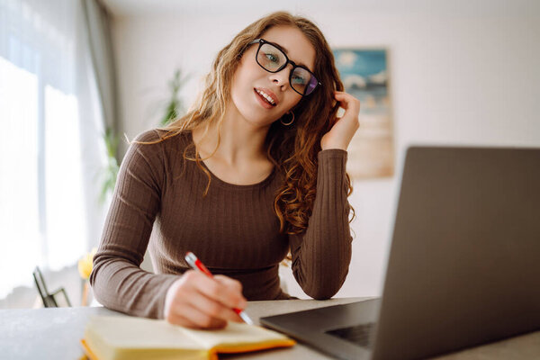 Beautiful businesswoman using a laptop wearing glasses while sitting at her workplace at home. Young woman working at home with a laptop. Concept of remote work, freelancing.