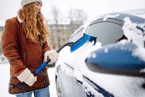 stock image Cleaning snow from a car. A beautiful woman is cleaning snow from a car with a brush. Transport concept, seasonality Winter glass cleaning.