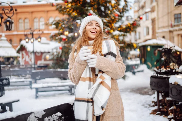 Genç bir kadın Noel pazarında sıcak bir içecek içiyor. Akşamları festival ışıklarıyla süsleniyor. Tatil konsepti, hafta sonları, rahatlama..